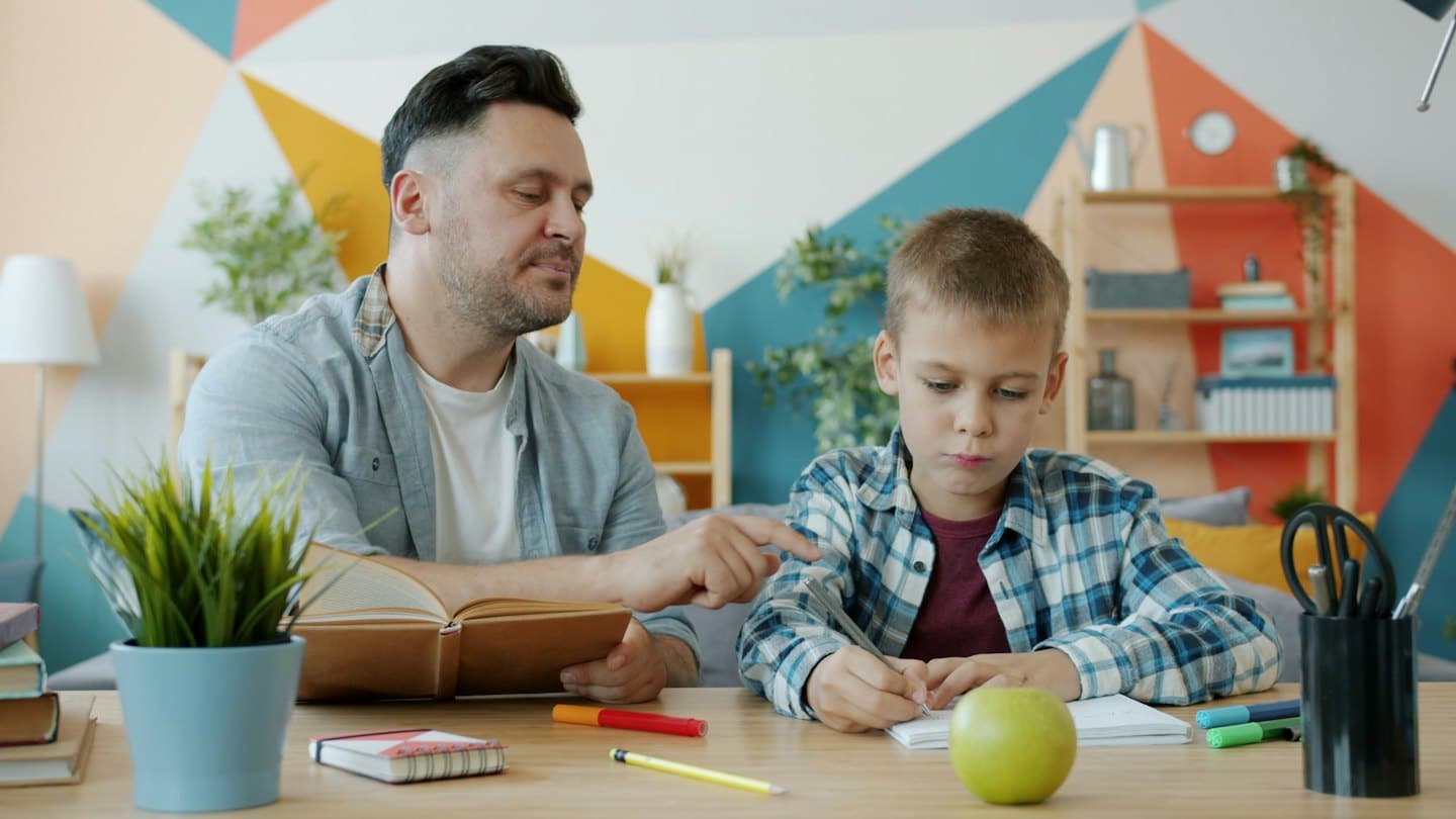 Mann hilft Junge beim Lernen an einem Schreibtisch mit bunter Wand.