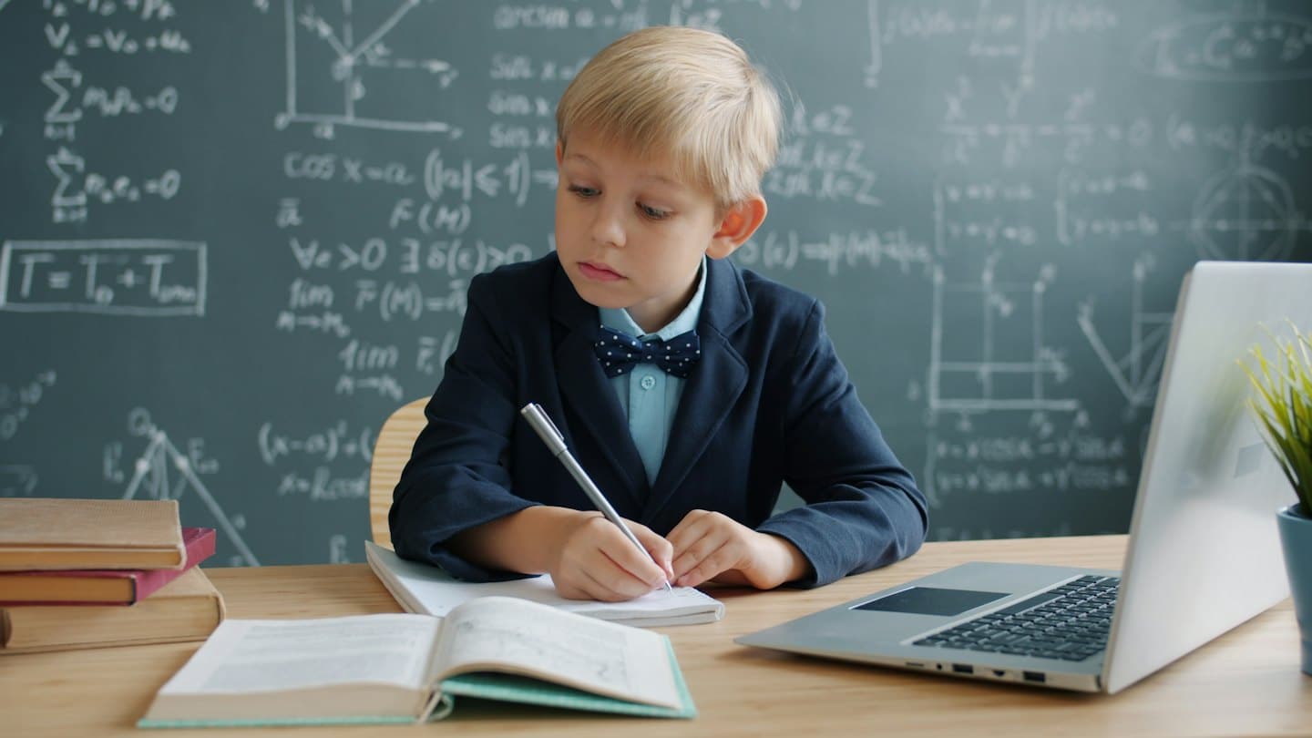 Ein Junge im Sakko schreibt an seinem Schreibtisch in ein Notizbuch.